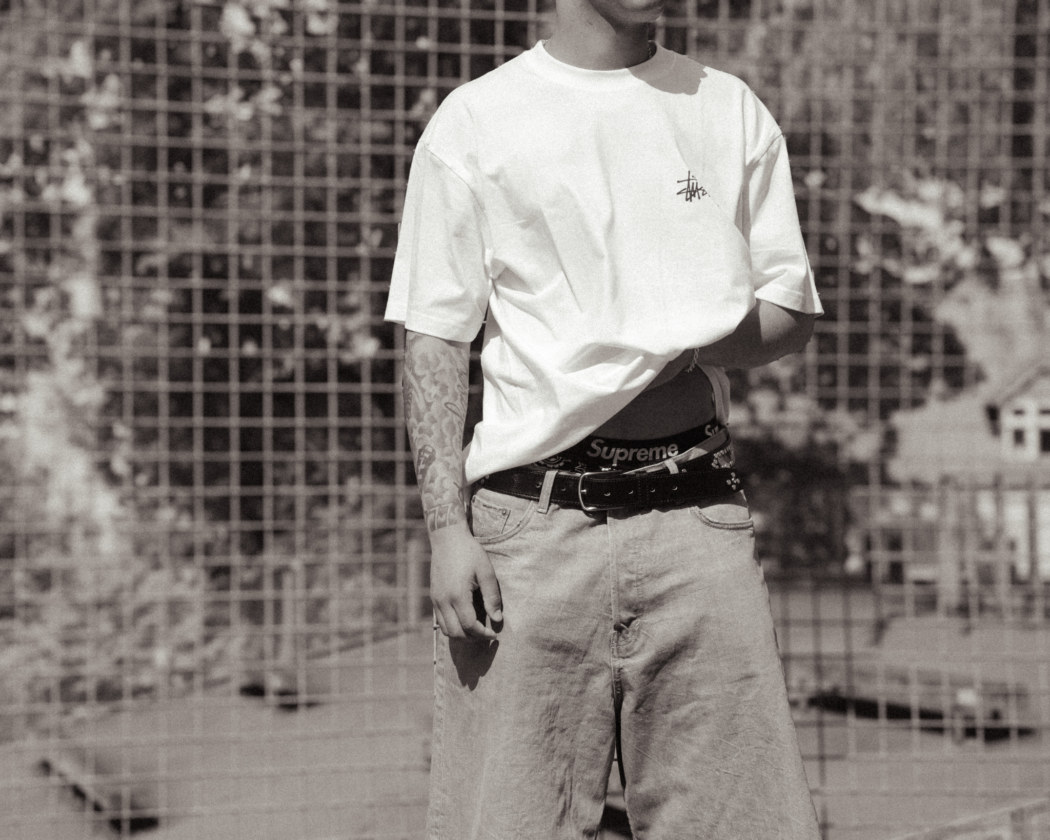 Model i hvid T-shirt, lyse jeans og Supreme-bælte foran et gitterhegn